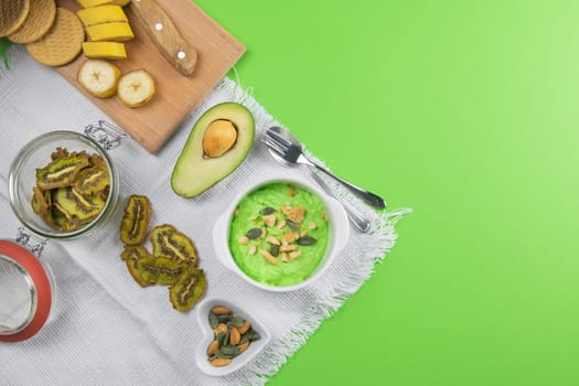 Two curd desserts: one with blueberries and nuts and the other with avocado and kiwi, next to there are pumpkin seeds and nuts.