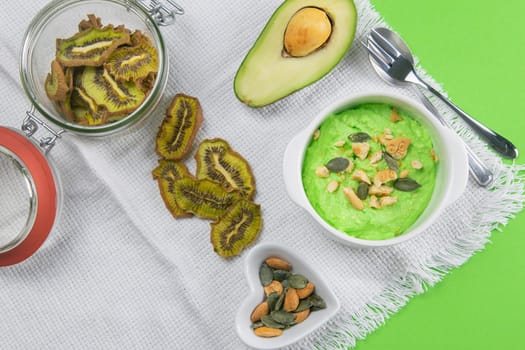 Two curd desserts: one with blueberries and nuts and the other with avocado and kiwi, next to there are pumpkin seeds and nuts.