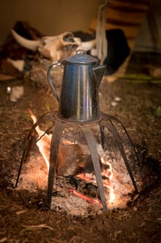 Old metal coffee pot stands over a campfire on a rack with cow skull