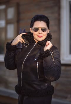 Brunette woman in a brown jacket and sunglasses holding a collar with hands, posing in front of the camera