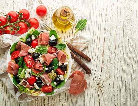 Healthy green salad with prosciutto, olives, tomato and fresh cheese on wooden table background. Healthy meal.