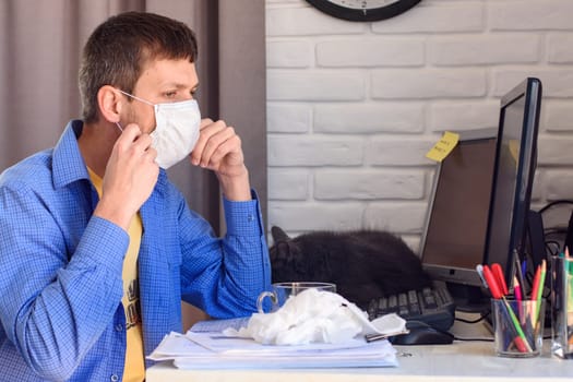 A man puts on a protective medical mask while working at home at the computer