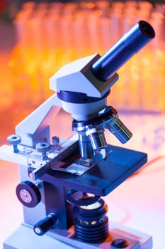 Close up of microscope lenses focused on a specimen in warm orange light light. Laboratory flask in the background.