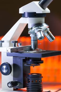 Close up of microscope lenses focused on a specimen in warm orange light light. Laboratory flask in the background.