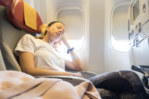 Tired blonde casual caucasian lady napping on uncomfortable seat while traveling by airplane. Commercial transportation by planes.