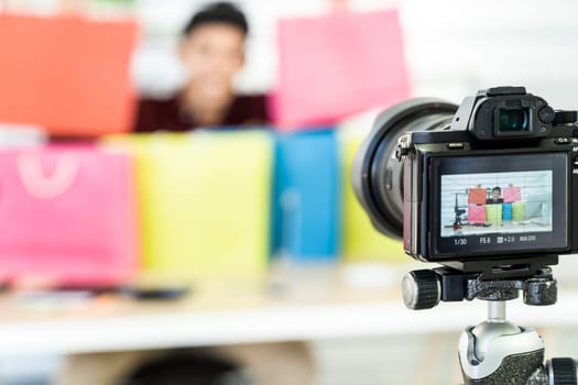 Young asian male online merchant blogger recording live vlog video with colorful shopping bag preparing for sale and review goods at home. Online influcencer on social media concept. Focus on camera