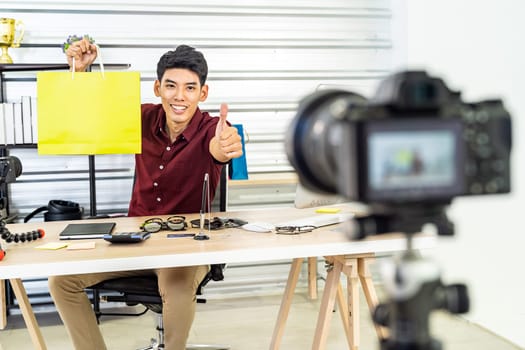 Young asian male online merchant blogger recording live vlog video with colorful shopping bag preparing for sale and review goods at home. Online influcencer on social media concept.