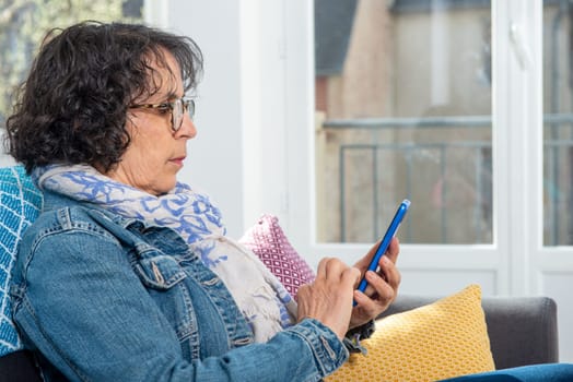 a cheerful brunette senior woman using smartphone while sitting on sofa