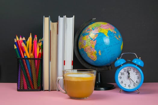 Coffe is in the foreground and behind him are a stack of books, a globe, a clock and a glass of pencils.