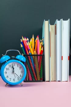 A stack of books, a clock and a glass with multi-colored pencils.