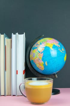 A stack of books, a globe and coffee with milk stand on a pink table.