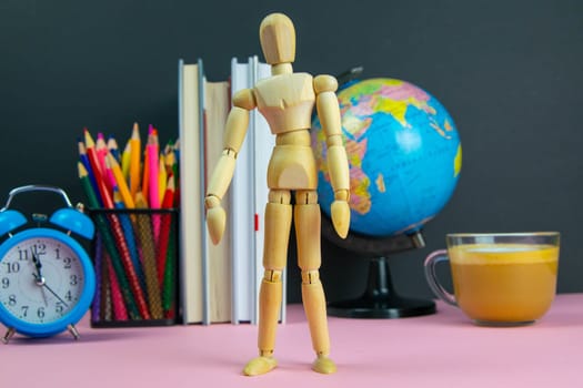 Figure of a wooden man is in the foreground and behind him are a stack of books, a globe, a clock and a glass of pencils.