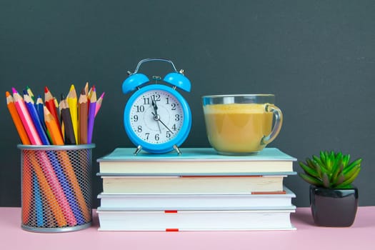A stack of books on which stands a coffe with milk next to stands a green flower and a glass with pencils.