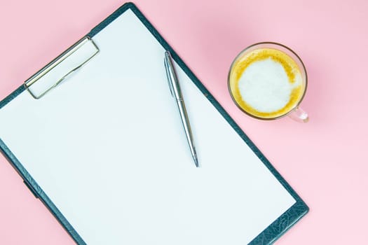 Tablet with a white sheet on which there is a silver pen and next to there is a glass with a coffe with milk.
