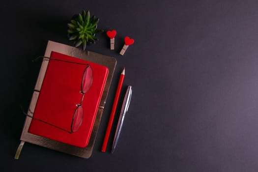 The red notebook stands on a gold notebook which stands on a black background, next to the notebook are a silver pen, a red pencil, two clothespins in the shape of a heart and glasses with a green plant.