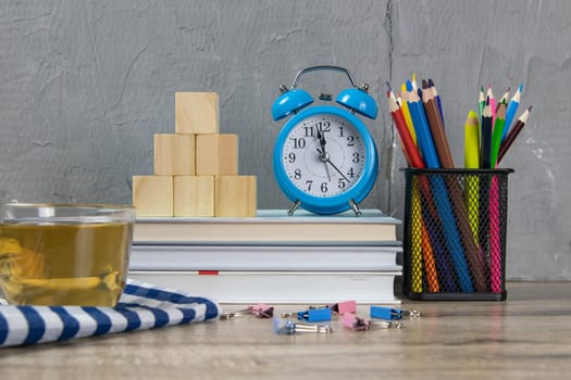 A stack of books, a clock, wooden cubes in the shape of a pyramid, next to there is tea with lemon in a transparent glass glass and multi-colored pencils.