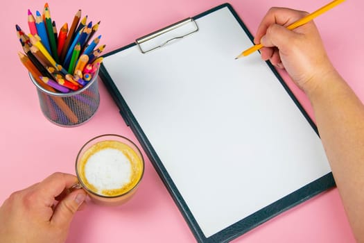 Tablet with white Leaf, next to which is a green plant, a glass with pencils. Hand holds a pen and writes a cup of coffee on the sheet in another hand