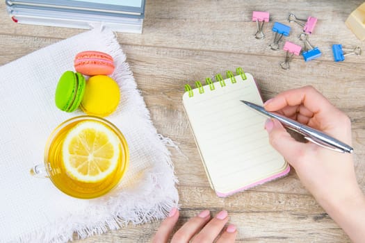 The hand holds a silver pen and writes in an open notebook, next to a notebook on a white towel there is mint tea with lemon and multi-colored muffins.