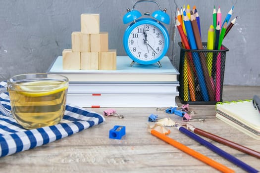 A stack of books, a clock, wooden cubes in the shape of a pyramid, next to there is tea with lemon in a transparent glass glass and multi-colored pencils.