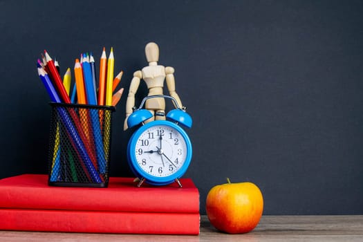 Two red books stand on the table, on the books there are blue watches and a glass with multi-colored pencils, behind the clock there is a figure of a wooden man and on the side of an apple. Front views with clear space