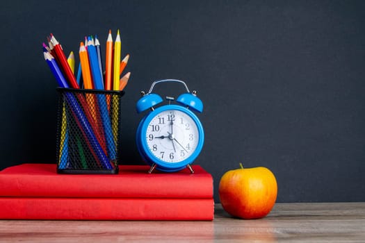 The blue alarm clock along with multi-colored pencils stand on a stack of red books, next to an apple.