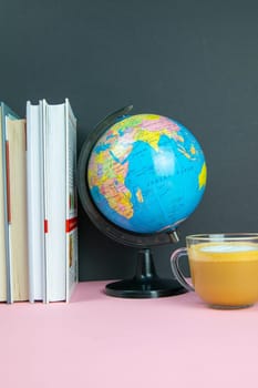 A stack of books, a globe and coffee with milk stand on a pink table.