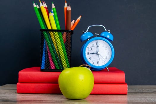 The blue alarm clock along with multi-colored pencils stand on a stack of red books, next to an apple.