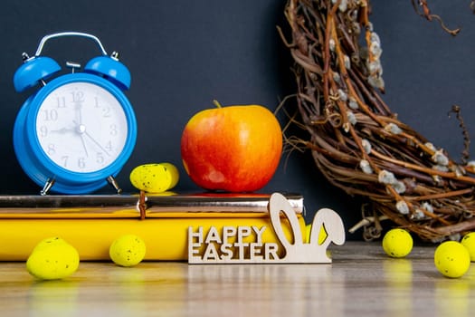 Easter concept, clock and apple stand on yellow books, eggs are scattered next to them, there is a wreath in the background.