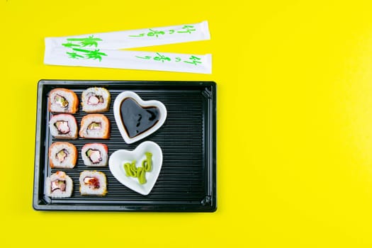 Black plate with sushi, filled with rolls of different kinds, next to stand sticks for sushi, red marinaginger and soy sauce on a yellow background. Top views with clear space