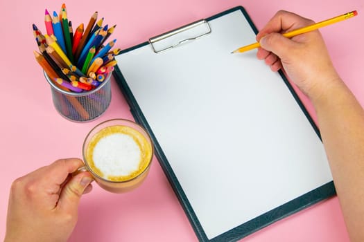 Tablet with white Leaf, next to which is a green plant, a glass with pencils. Hand holds a pen and writes a cup of coffee on the sheet in another hand