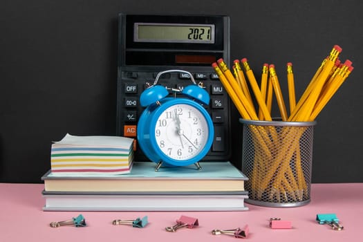 On a stack of books there is a calculator, a clock and sheets for notes, and next to a glass of pencils.