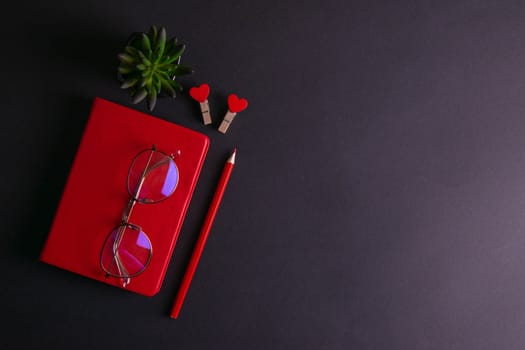 The red notebook stands on a black background, on the notebook there are glasses and next to the notebook, a flower, two clothespins in the shape of hearts and a red pencil.