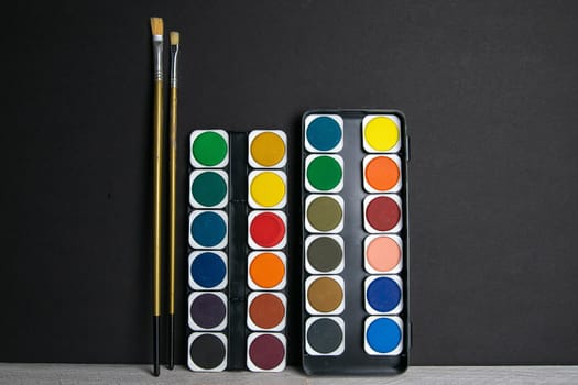 Colorful paints stand on a wooden table propped up against a black wall. Frontal view.