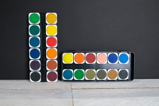 Colorful paints stand on a wooden table propped up against a black wall. Frontal view.