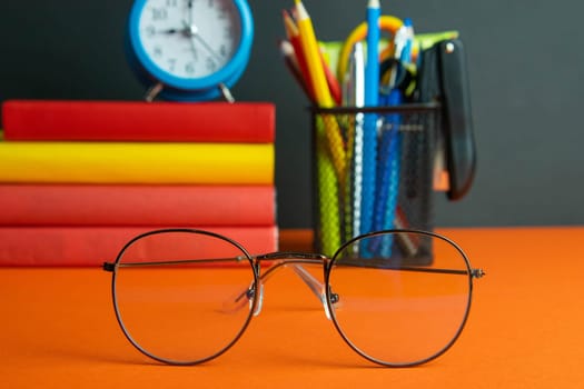 A stack of books, women's glasses in the foreground, blue clock and a glass of pencils stand on an orange background.