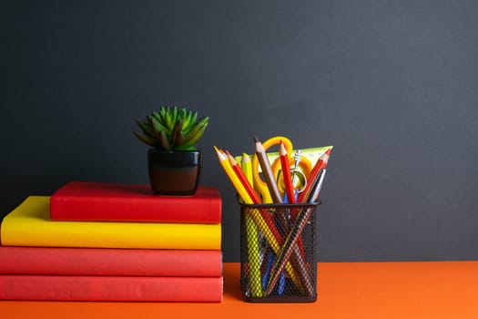 A stack of books, a green flower, and a glass of pencils stand on an orange background.