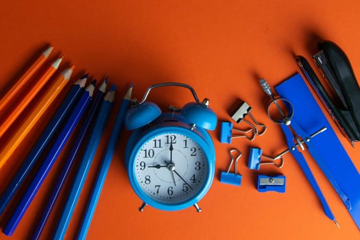Pencils, blue alarm clock, compass, ruler and stapler on an orange background.