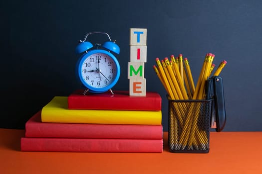 A stack of books, a green flower, blue clock and a glass of pencils, and wood cubes with text time stand on an orange background.