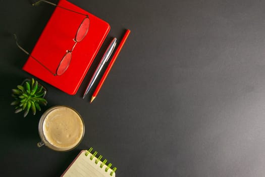 The red notebook stands on a black background, on the notebook there are glasses and next to the notebook, a flower, coffe, pen and a red pencil.