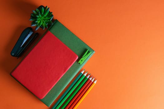 Red and green notebooks stand on an orange background, next to notebooks there are multi-colored pencils, and a green plant.