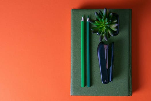 Green notebook stands on an orange background, on a notebook there are two green pencils, a green plant and a stapler.