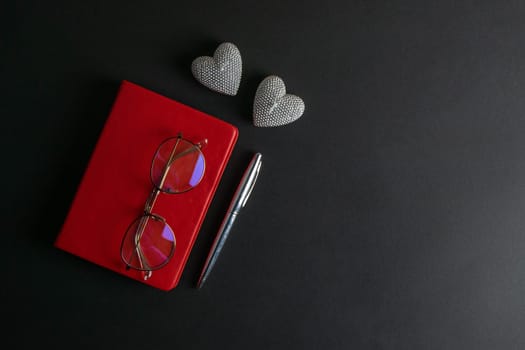 The glasses stand on a red notebook, there are two heart-shaped candles nearby, and a silver pen.
