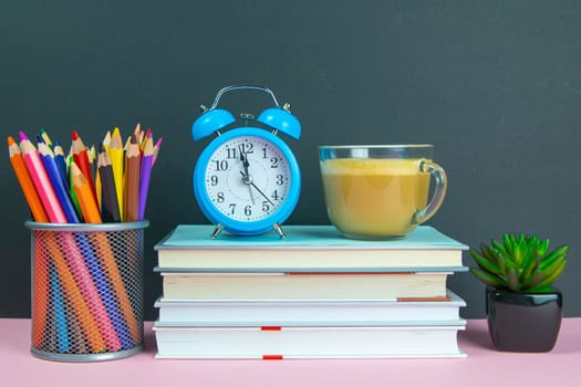 A stack of books on which stands a coffe with milk next to stands a green flower and a glass with pencils.