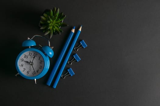 Blue alarm clock, green plant along with blue pencils and pins on a black background.