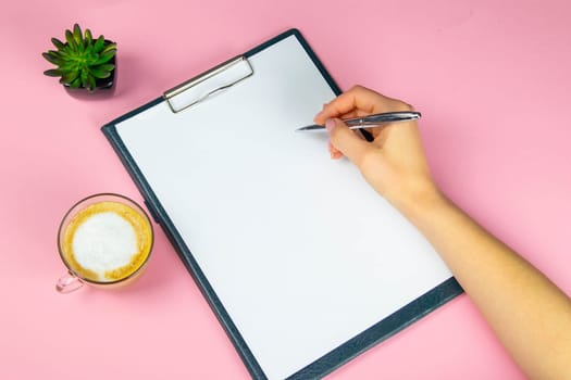 Tablet with white flattery next to which stands a green plant, hand holds a pen and writes on a sheet in the other hand a cup with a coffe.