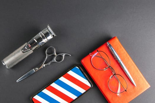 A machine for haircuts, scissors, comb stand on a white towel and next to there is a Phone, pen and a red notebook on which there are women's glasses.
