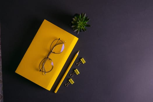 Yellow pencil, yellow notebook and glasses, pins, clip paper on on black background.