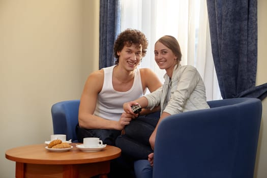 Happy young couple with breakfast are sitting in armchairs and watching TV in living room at home.