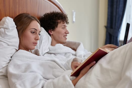 Young family with laptop and book are lying on bed in room at home. Weekend or isolation period, social distancing. Remote learning and work. Happy young couple having a rest. Selective focus on woman