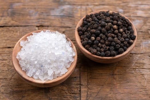 Coarse crystal salt and whole black peppercorns in small wooden bowls on a rustic wooden background
